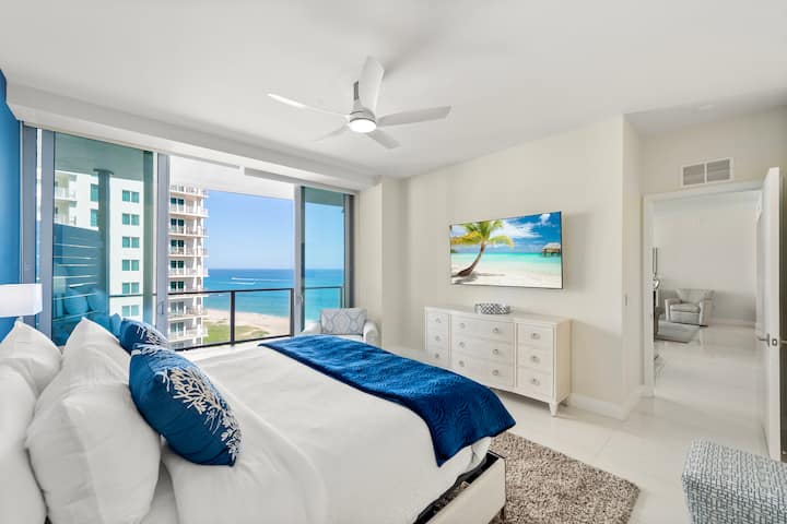 The king suite dressed in white and navy, with a ceiling fan and the ocean through the corner glass