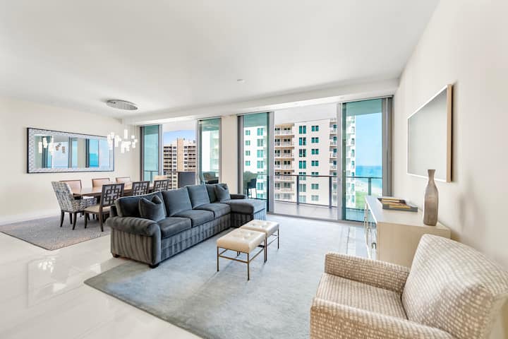 The open living and dining room wrapped in glass on two sides, with a sectional sofa facing the ocean